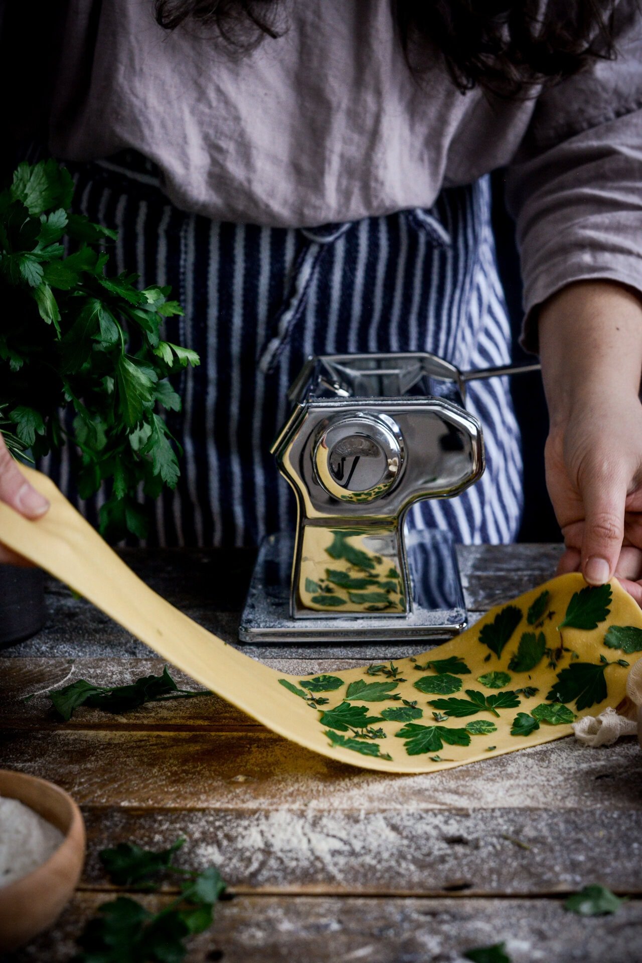 A person uses a pasta machine to roll out sheet pasta with fresh parsley leaves embedded in the dough. Flour is scattered on a wooden surface, and a bunch of parsley is nearby.