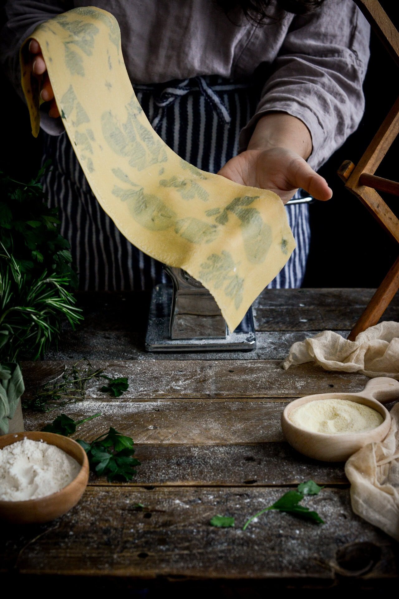 A person wearing an apron feeds a sheet of homemade herb pasta through a pasta machine. Fresh herbs, flour, and semolina are on a rustic wooden table nearby.