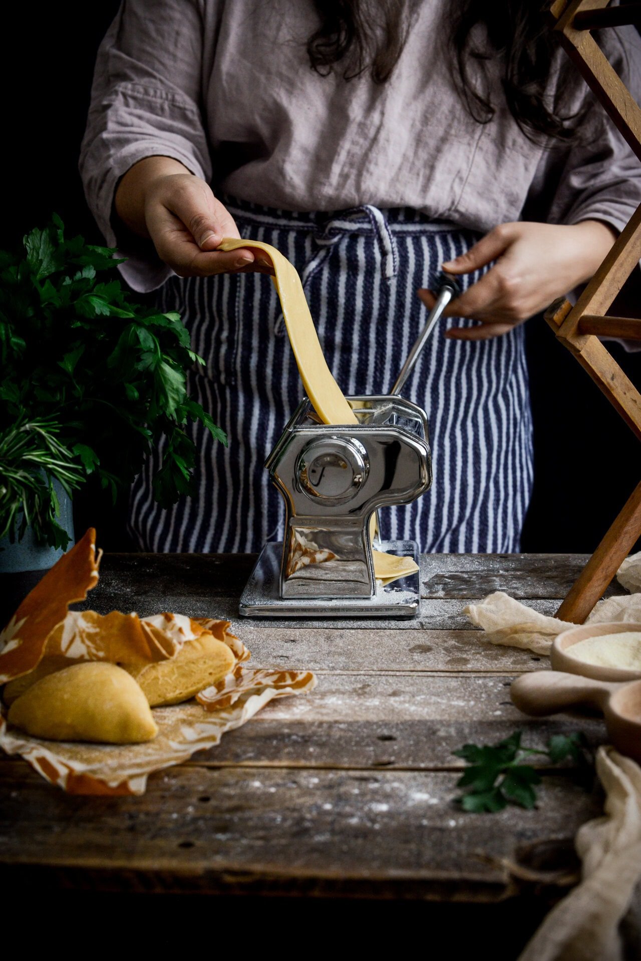 A person in a striped apron uses a pasta machine to roll out fresh pasta dough on a rustic wooden table, surrounded by ingredients and tools for homemade pasta.