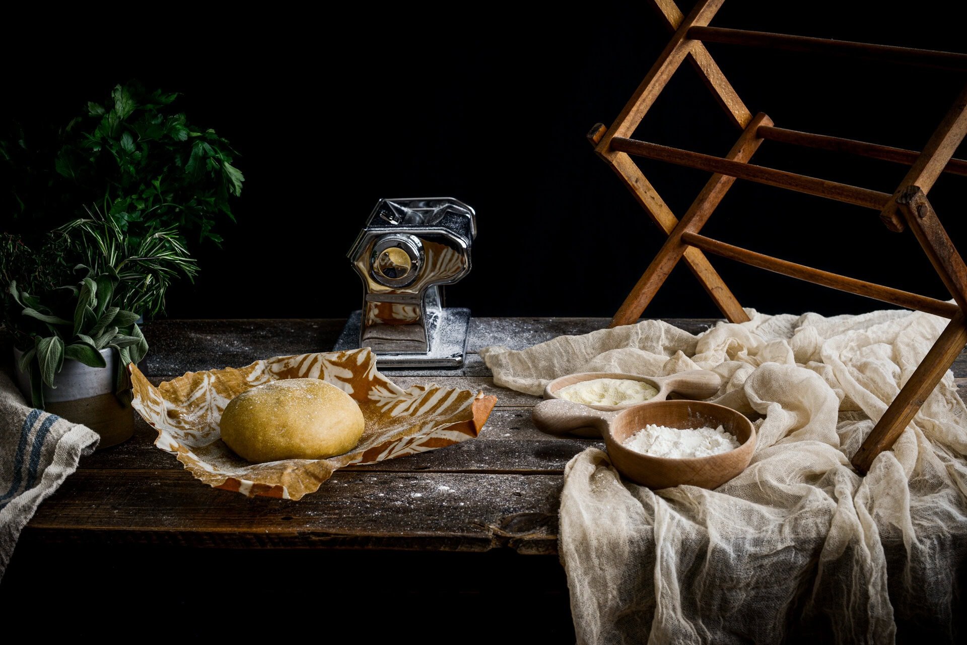 A ball of pappardelle pasta dough on parchment paper sits on a rustic wooden table with bowls of flour and cheese, fresh herbs, a pasta machine, and a wooden drying rack under soft, moody lighting.