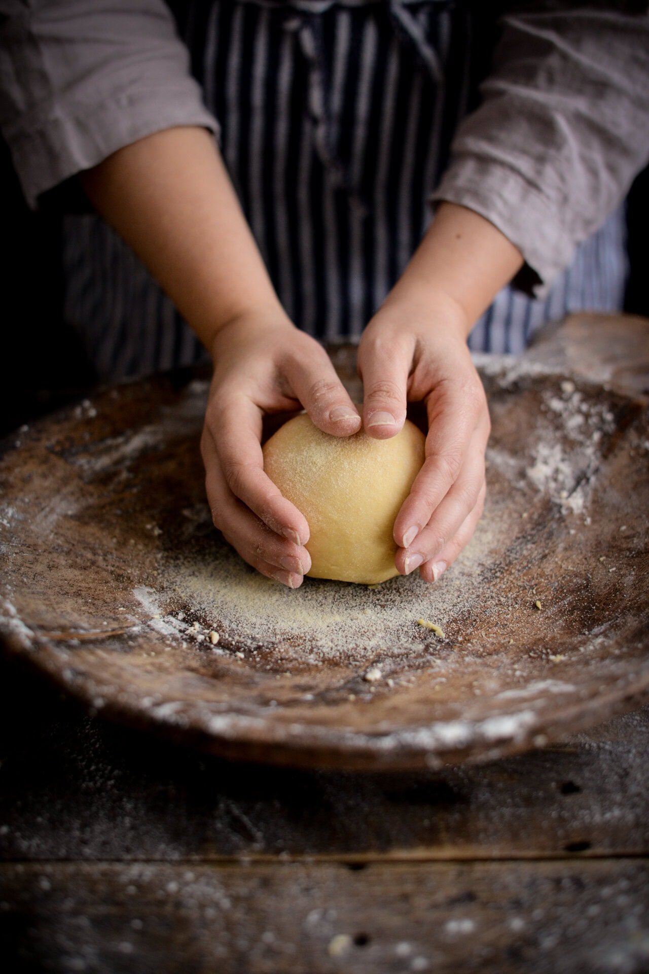 A person shaping dough with their hands on a rustic wooden surface dusted with flour, wearing a striped apron and a light long-sleeved shirt.