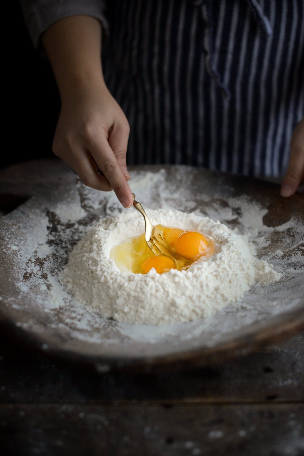 A person in an apron uses a fork to mix two cracked eggs in a well of flour on a wooden surface, preparing dough.