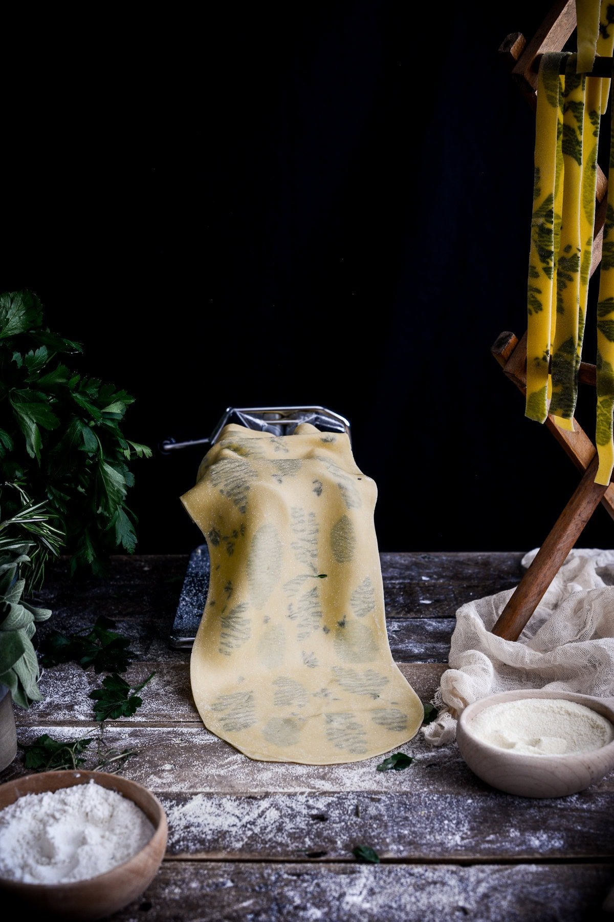 A sheet of uncooked pasta with herbs is draped over a pasta machine on a rustic wooden table, surrounded by flour, a bowl of semolina, fresh herbs, and a pasta drying rack.