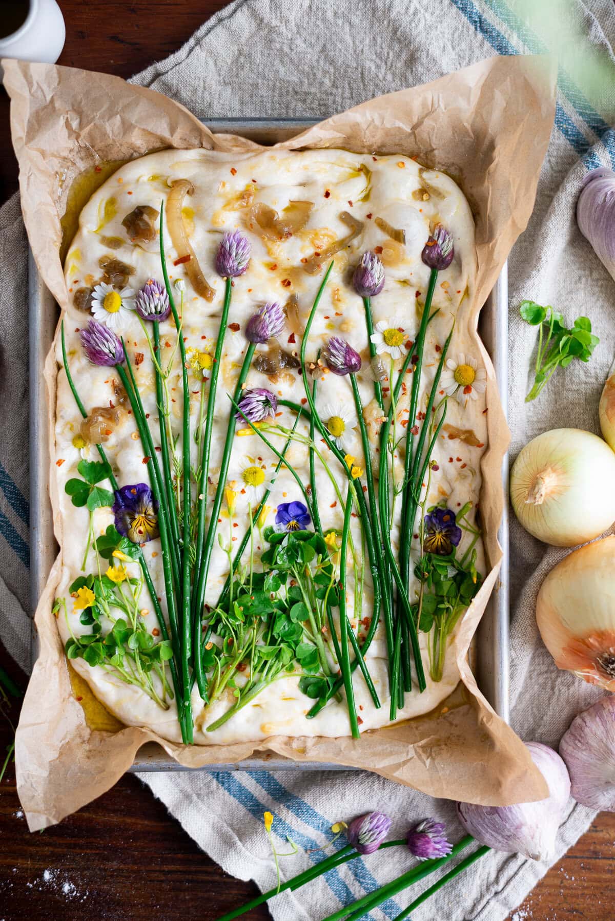 A rectangular sourdough focaccia decorated with chive flowers, chive stems, and edible blooms to resemble a garden, on parchment in a baking tray. Onions, garlic, and herbs are scattered around on a rustic cloth.