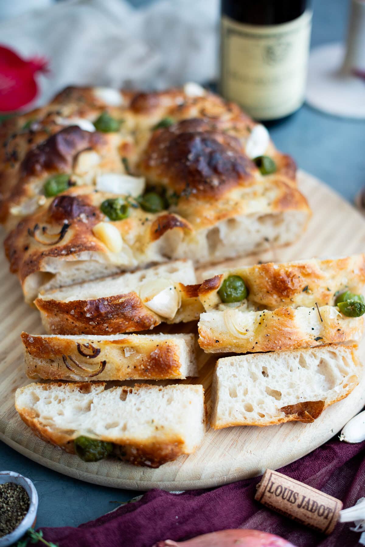 A round loaf of sourdough focaccia topped with onions, garlic cloves, and green olives, partially sliced on a wooden board. A wine bottle, cork, and small bowl of pepper are nearby—perfect for trying your next sourdough focaccia recipe.