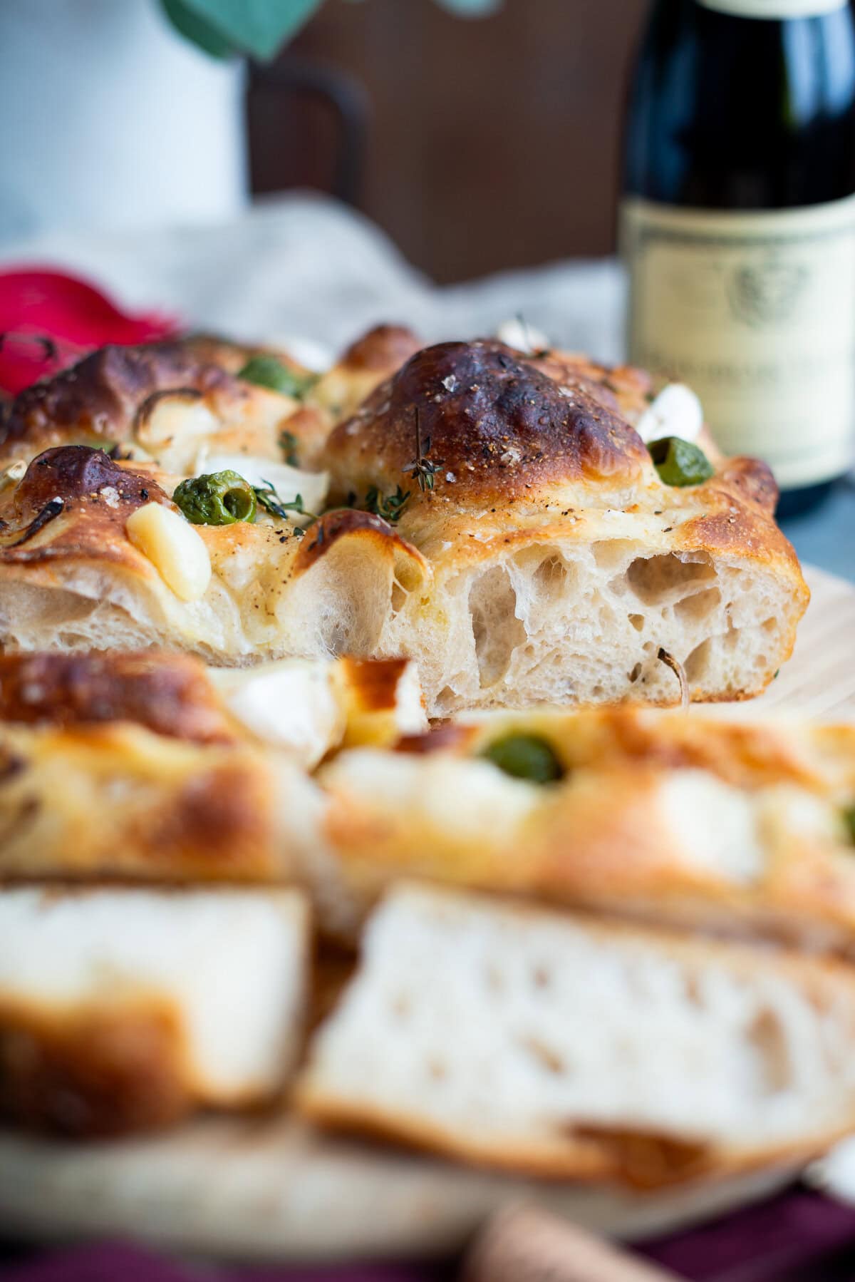 A close-up of freshly baked sourdough focaccia topped with herbs and garlic, with a bottle of wine in the background. The bread looks airy and golden brown with a crisp crust, perfect for trying a new sourdough focaccia recipe.