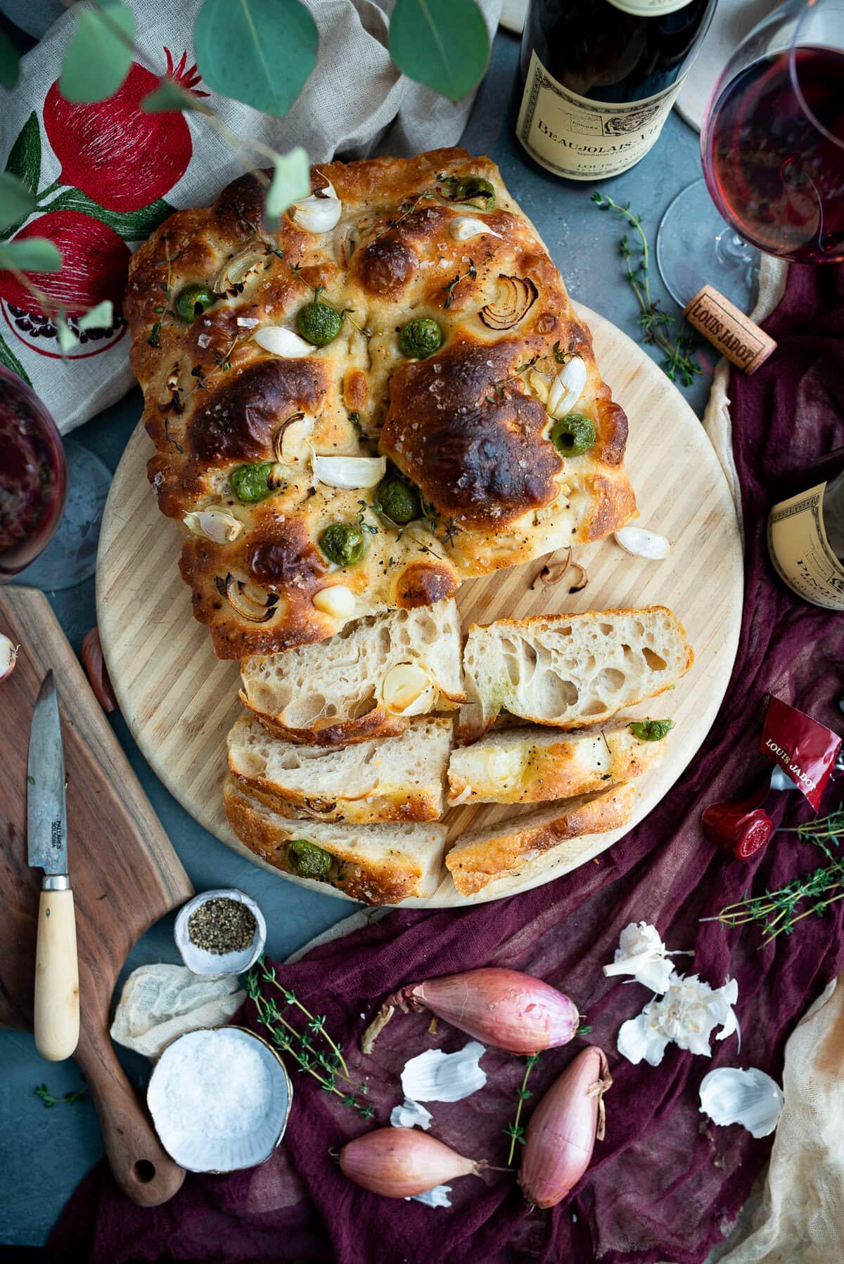 A round wooden board holds sliced sourdough focaccia topped with green olives, garlic, and herbs, surrounded by wine glasses, bottles, shallots, salt, pepper, a knife, and fresh thyme on a rustic table with a burgundy cloth.
