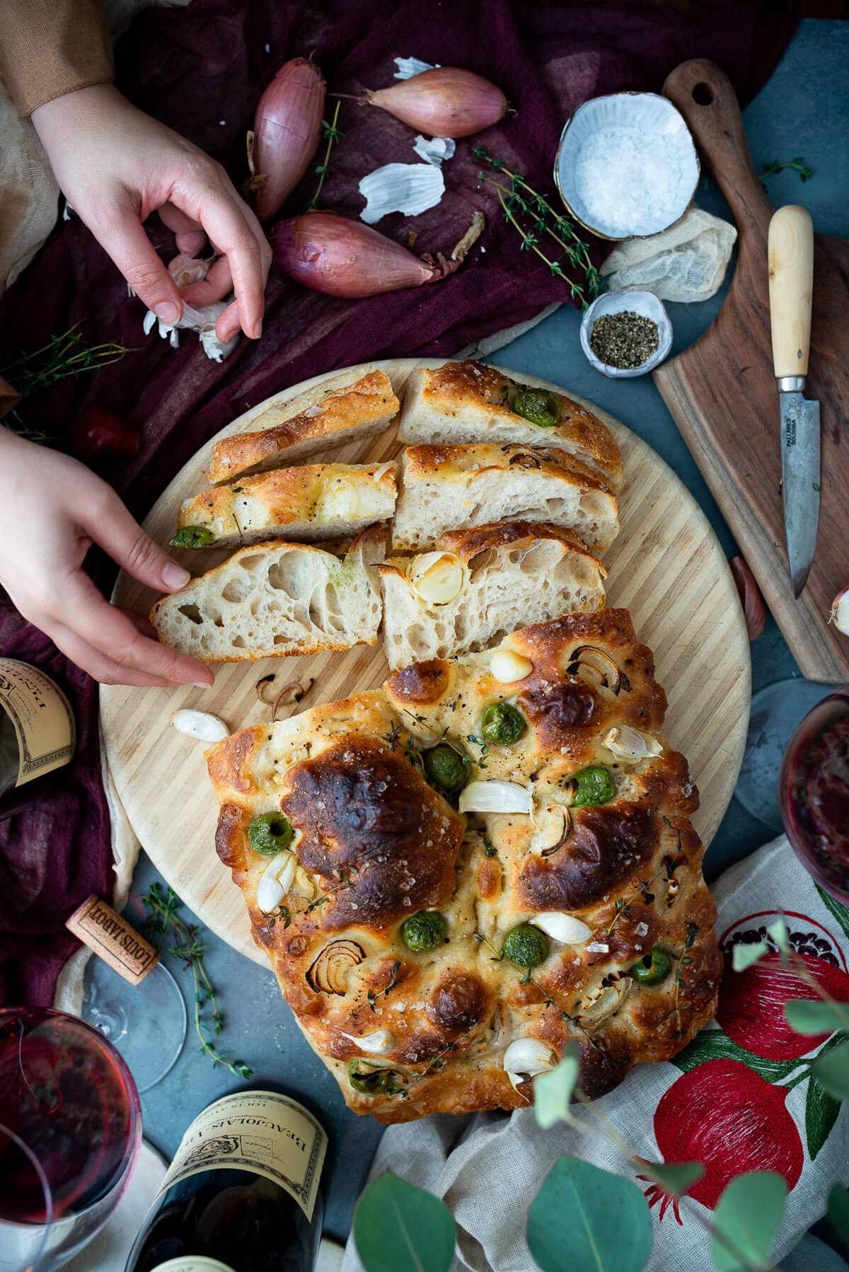 A hand reaches for a slice of sourdough focaccia on a round wooden board, surrounded by wine bottles, shallots, salt, pepper, and herbs atop a blue tabletop with a burgundy cloth.