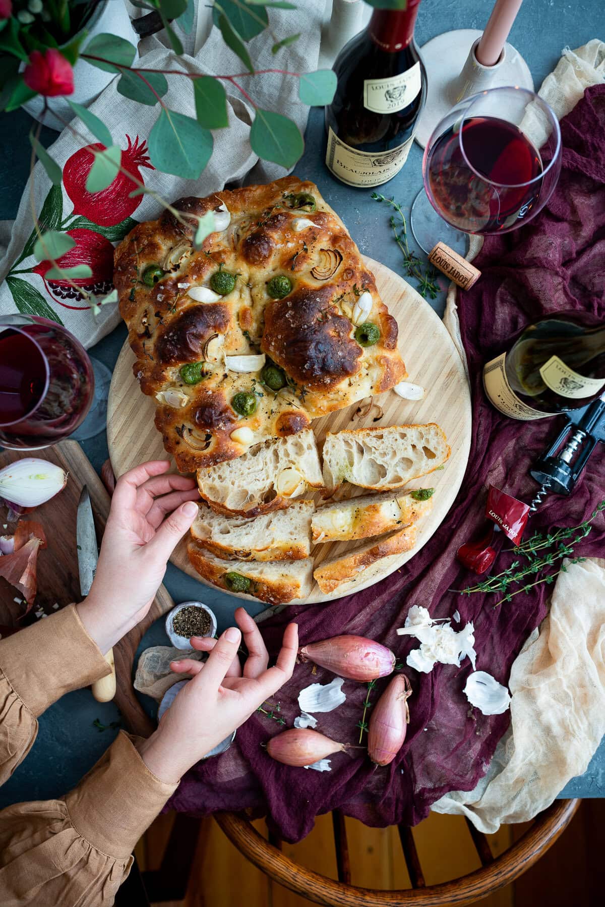 A person slices freshly baked sourdough focaccia topped with herbs and vegetables on a wooden board, surrounded by wine bottles, filled wine glasses, shallots, garlic, and decorative elements on a rustic table.