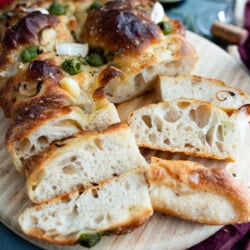 A sliced loaf of homemade sourdough focaccia bread on a wooden board, featuring a golden, bubbly crust and airy interior. Garlic cloves and herbs are baked on top, with a glass of red wine and a bottle in the background.