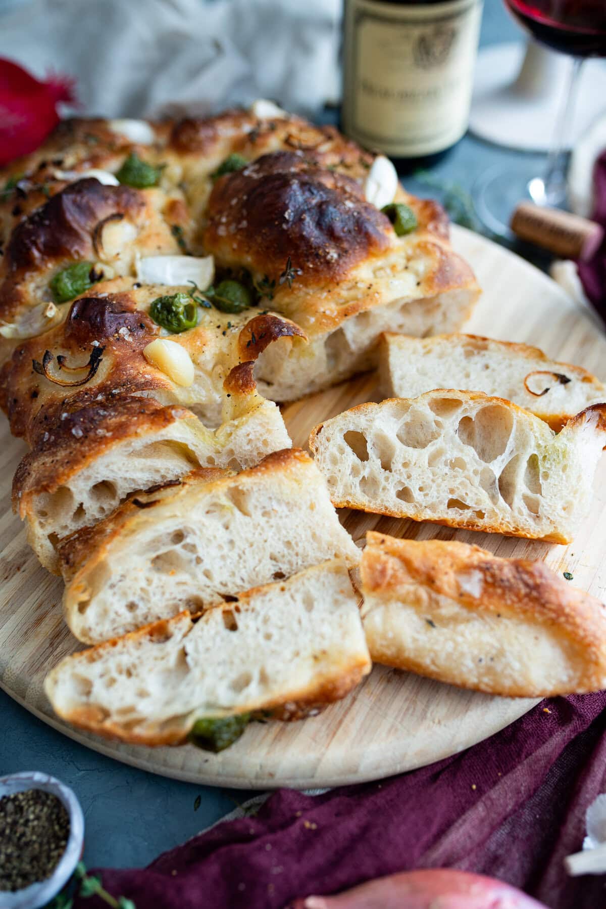 A sliced loaf of homemade sourdough focaccia with a golden, crispy crust, topped with herbs, garlic, and green chilies, displayed on a wooden board alongside a glass of red wine and a wine bottle in the background.
