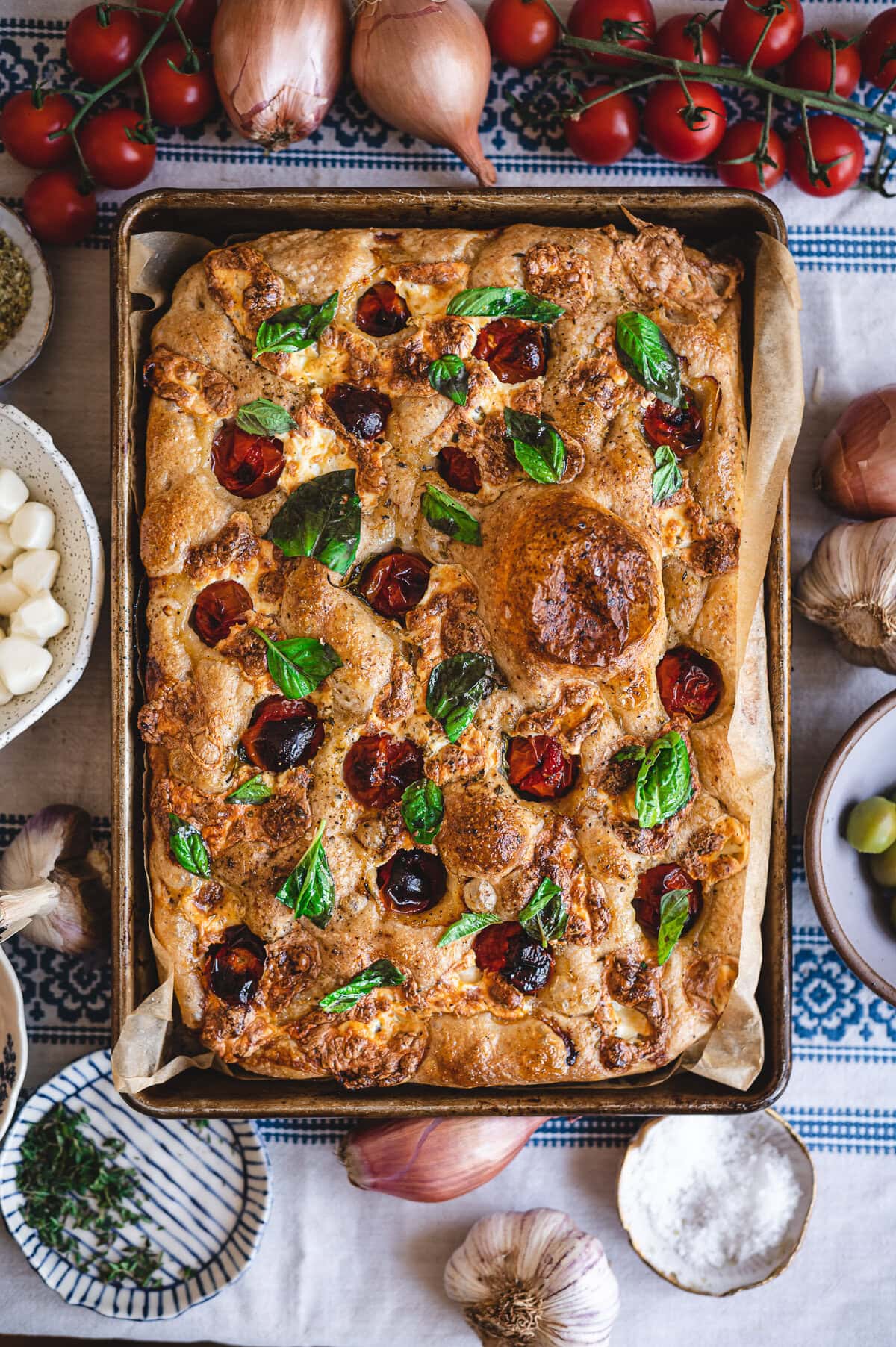 A baked sourdough focaccia topped with cherry tomatoes, fresh basil, and bubbling cheese sits on parchment in a tray, surrounded by garlic, shallots, herbs, and tomatoes on a patterned cloth. Try this delicious sourdough focaccia recipe today!.