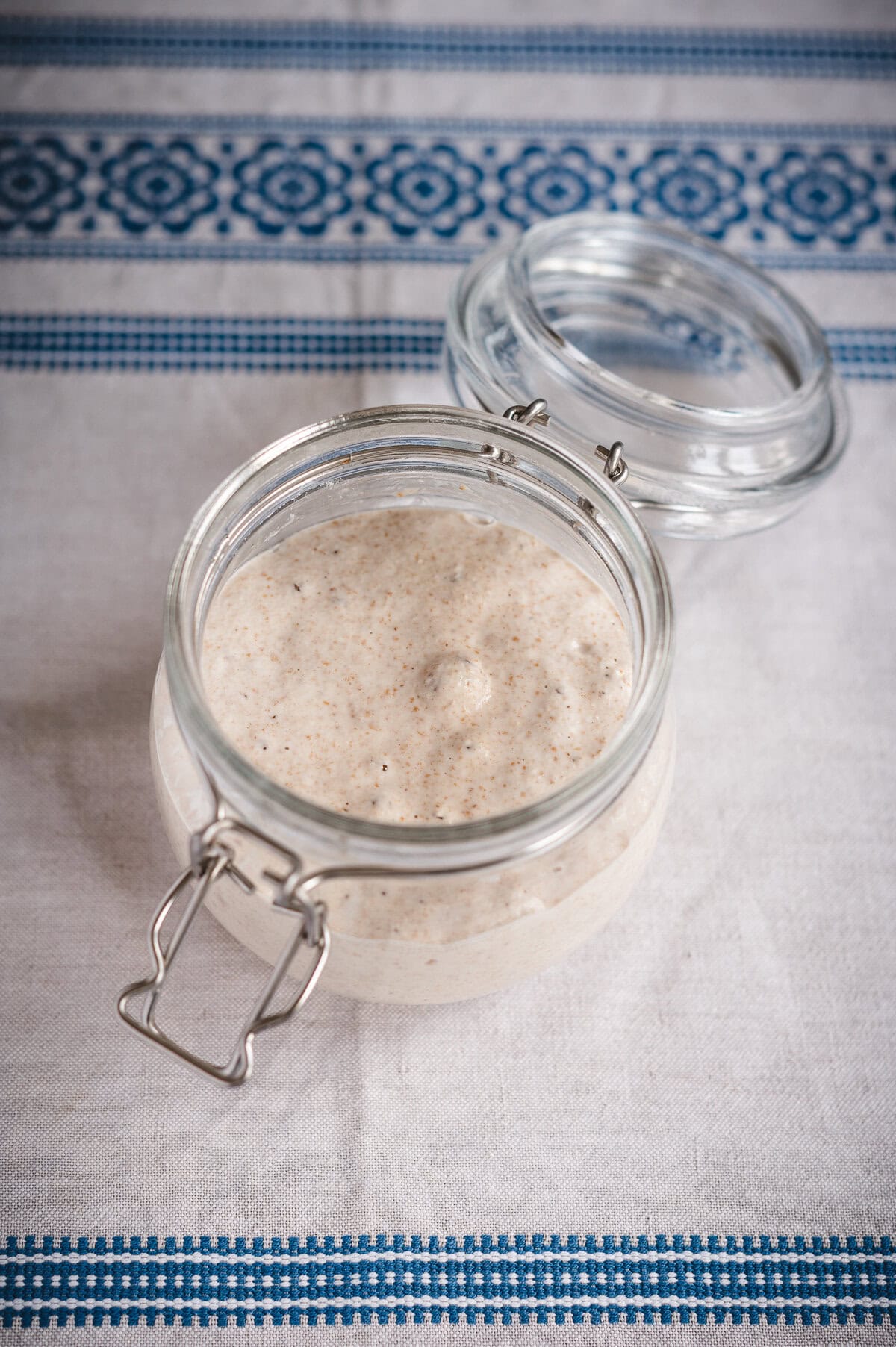 An open glass jar filled with creamy, beige sourdough starter sits on a white tablecloth with blue decorative patterns. The jar’s hinged lid rests to the side, ready for your next sourdough focaccia recipe.