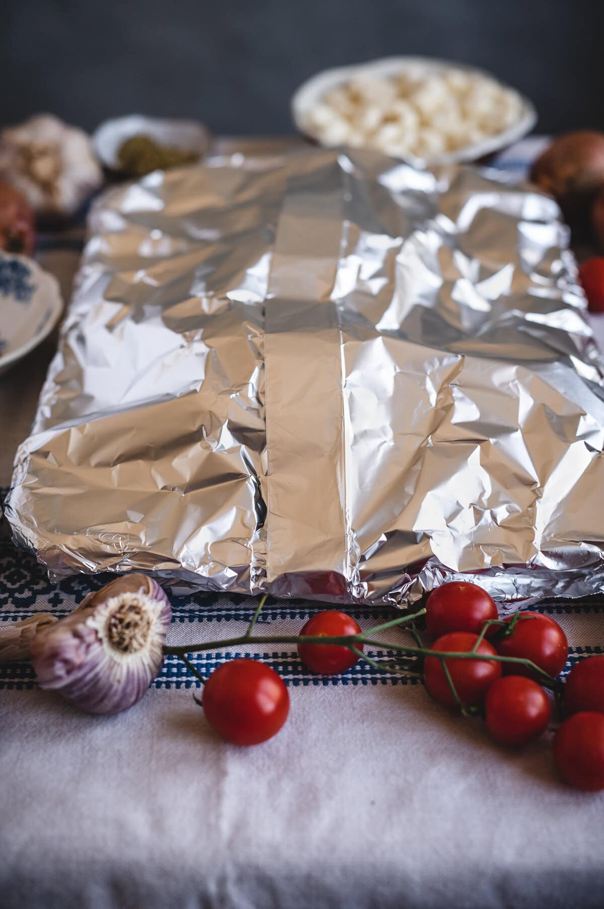 A baking dish covered with aluminum foil sits on a table surrounded by fresh cherry tomatoes, a garlic bulb, and plates of chopped vegetables—perfect accompaniments for a homemade sourdough focaccia recipe.