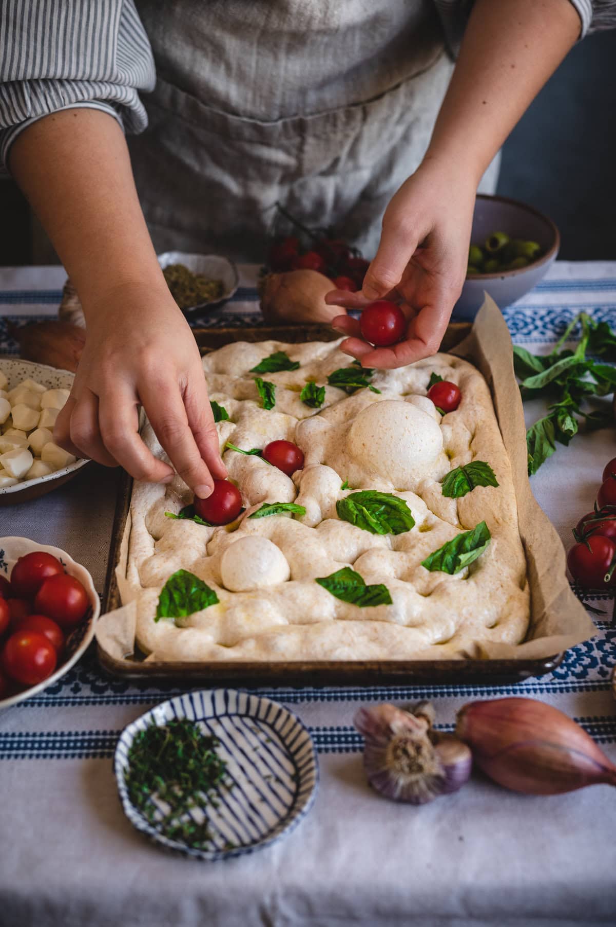 A person places cherry tomatoes and basil leaves on unbaked sourdough focaccia dough in a baking tray, surrounded by cheese, herbs, and garlic—preparing for a fresh and flavorful sourdough focaccia recipe.