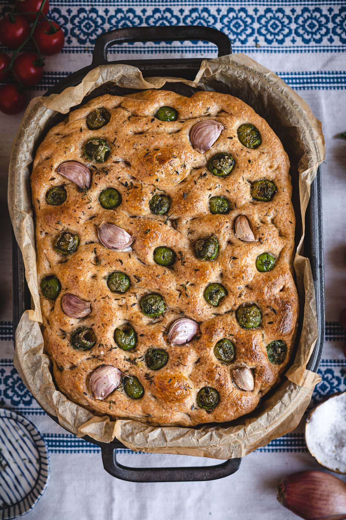 Golden-brown sourdough focaccia in a rectangular pan, topped with whole garlic cloves and sliced green olives on parchment paper. The background features a blue and white patterned cloth. Try this delicious sourdough focaccia recipe today!.