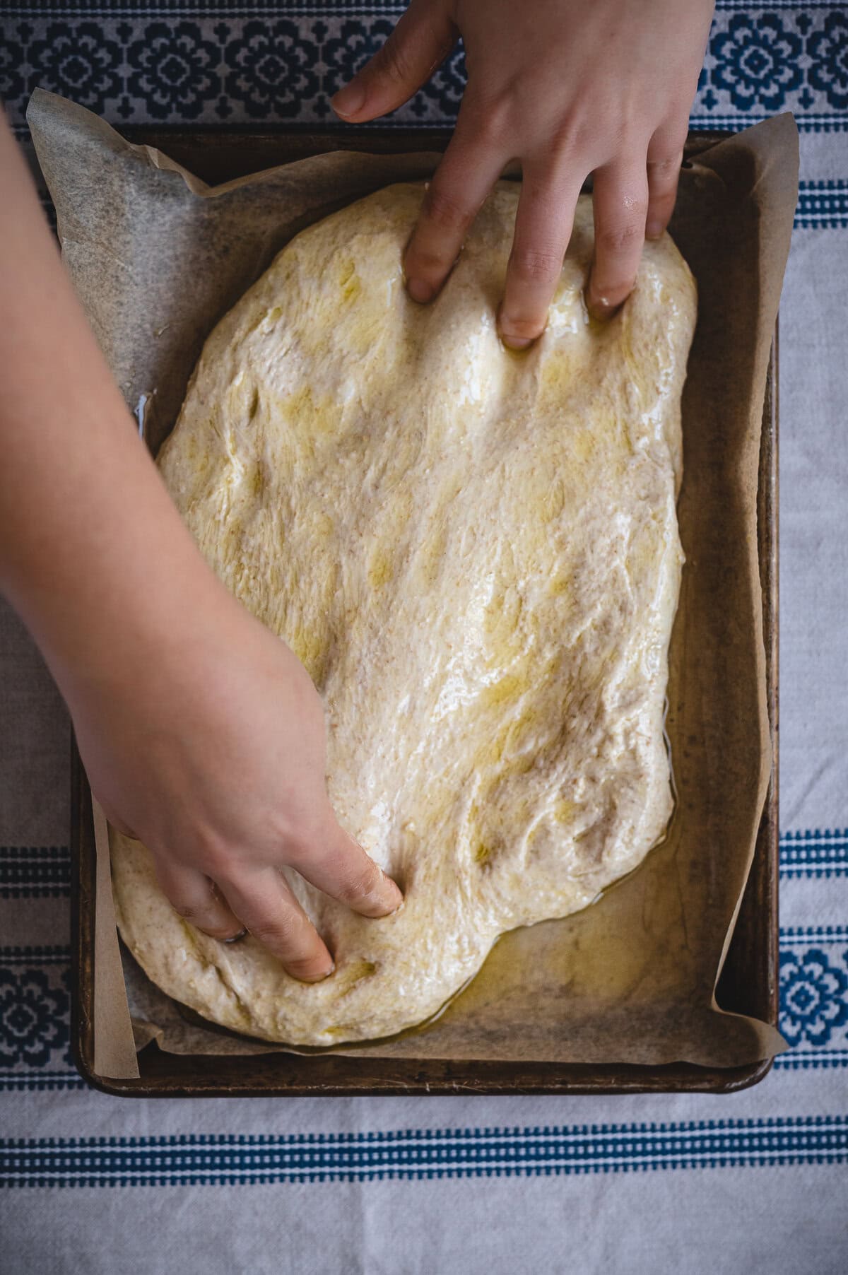 A person’s hands are stretching and pressing sourdough focaccia dough into a rectangular baking pan lined with parchment paper, preparing it for baking. The dough appears to be brushed with oil and is on a patterned cloth.