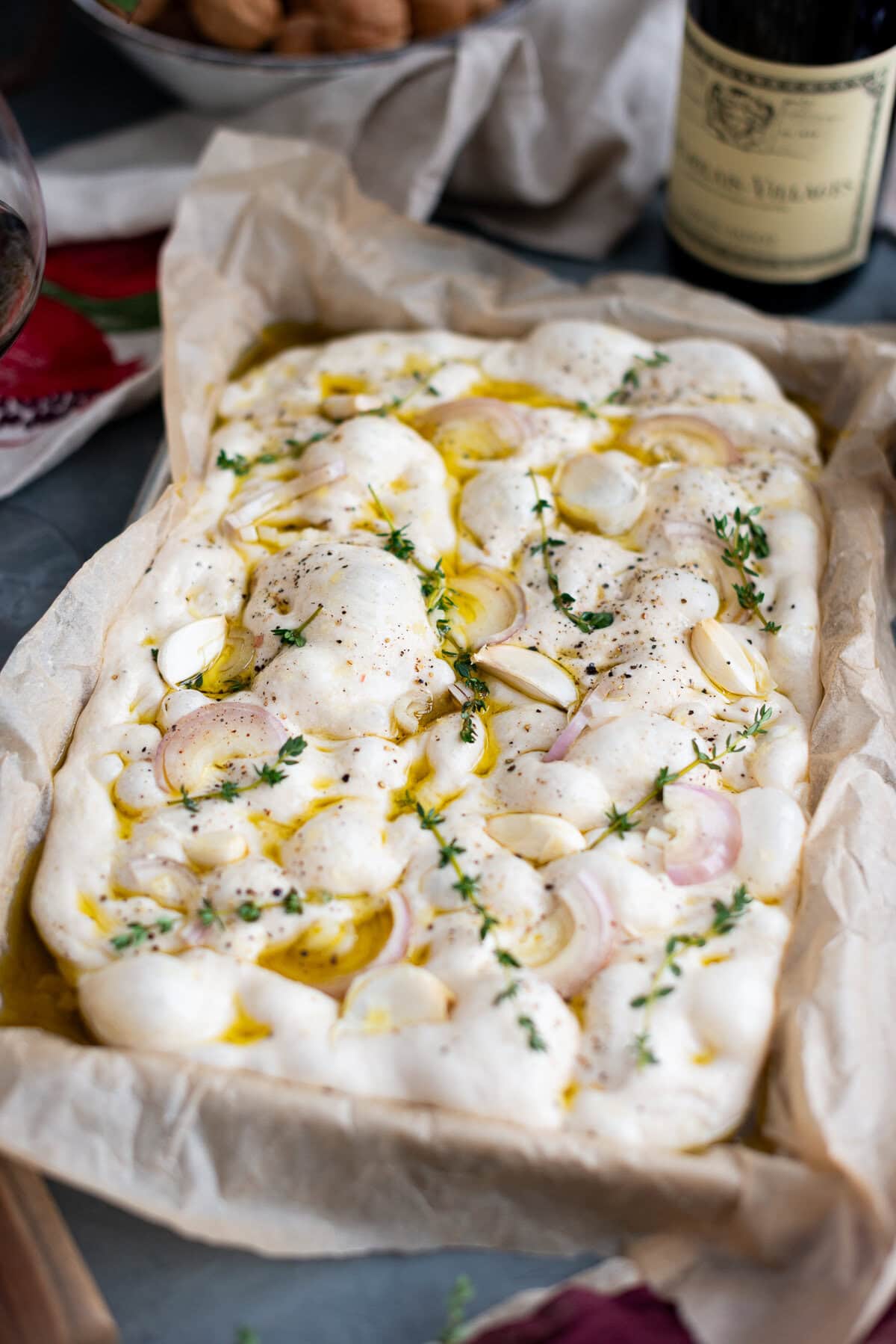 A rectangular sourdough focaccia dough topped with sliced garlic, shallots, fresh thyme, olive oil, and cracked pepper sits on parchment paper, ready to bake. A bottle of wine and walnuts are in the blurred background.
