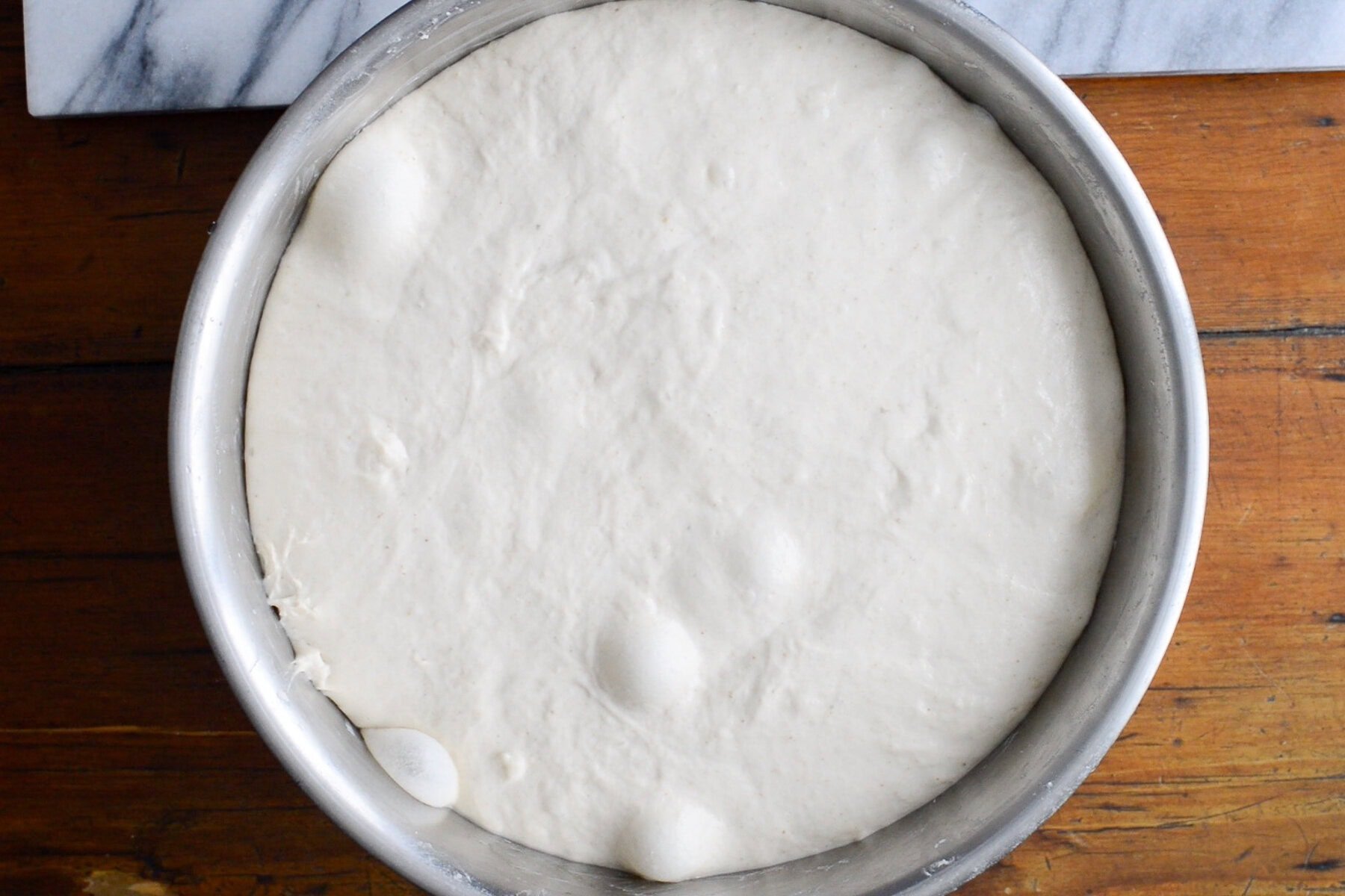 A large metal bowl filled with risen pizza dough, showing bubbles on the smooth, pale surface, sits on a wooden table next to a marble board—perfect for starting a sourdough focaccia recipe.
