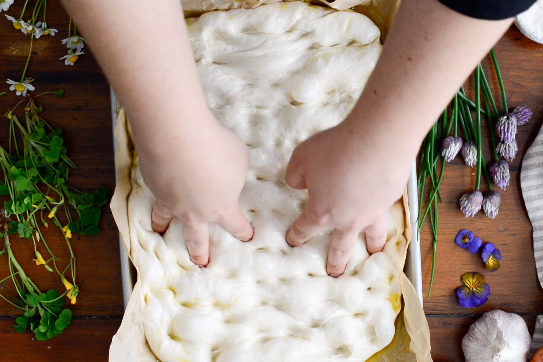 Two hands pressing fingertips into a rectangular tray of sourdough focaccia dough. The scene is surrounded by herbs and flowers on a wooden table, suggesting a rustic, homemade sourdough focaccia recipe in the making.