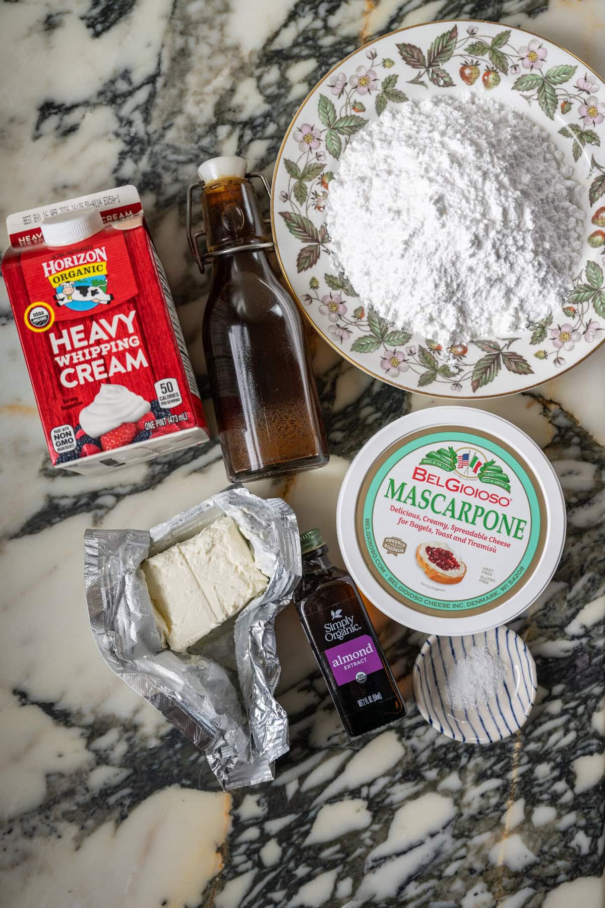 A marble countertop displays berry chantilly cake frosting ingredients: heavy whipping cream, vanilla, powdered sugar, mascarpone, cream cheese, almond extract, and salt.