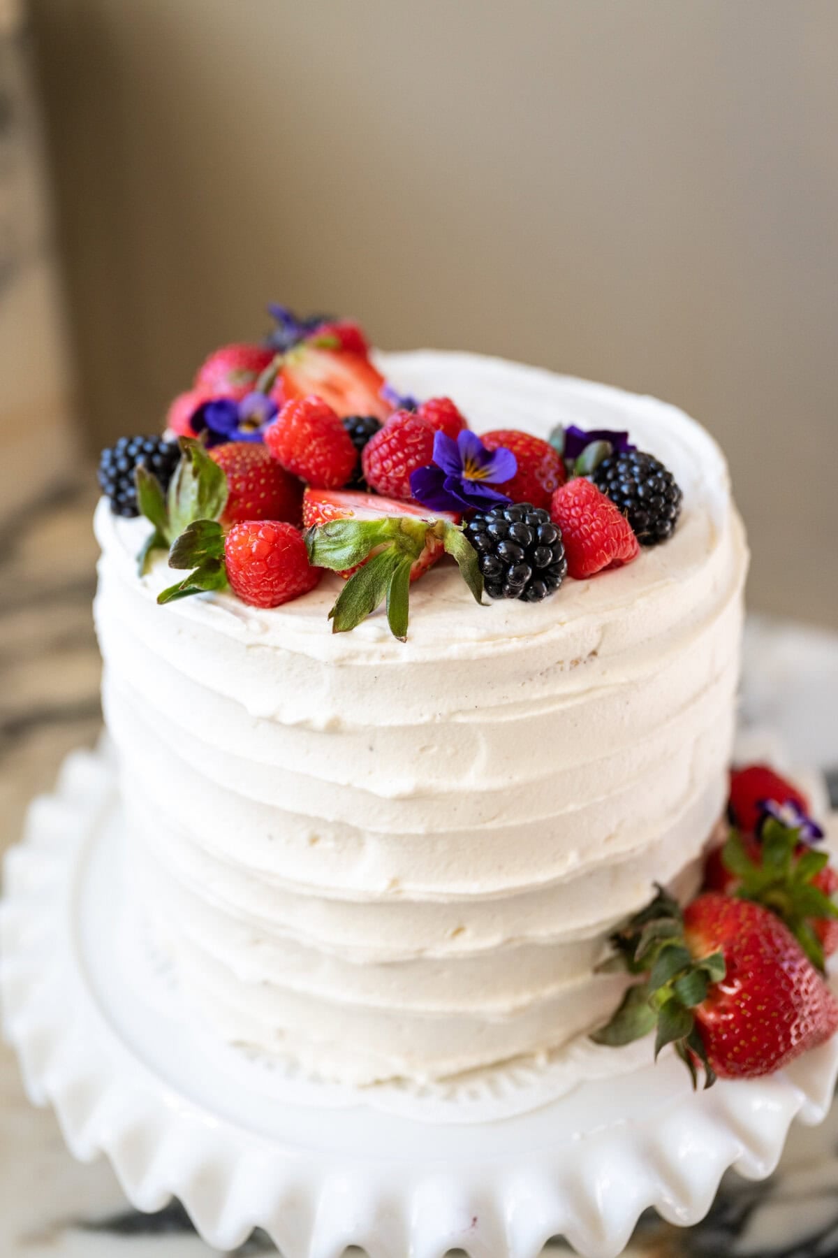 A white frosted berry chantilly cake topped with fresh strawberries, raspberries, blackberries, and purple edible flowers, displayed on a white cake stand.