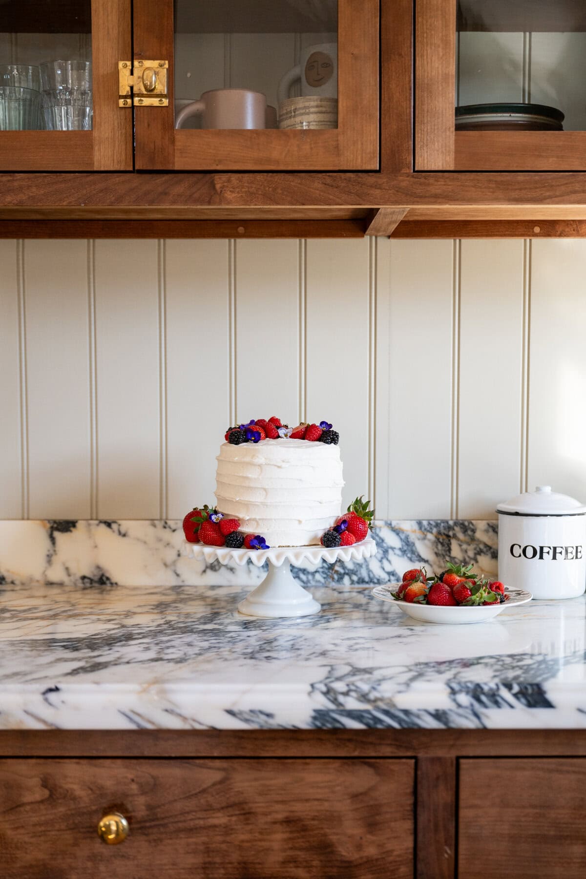 A Whole Foods copycat berry chantilly layer cake, sits on a cake stand on a marble counter, topped with fresh berries and surrounded by strawberries and a coffee canister under wooden cabinets.