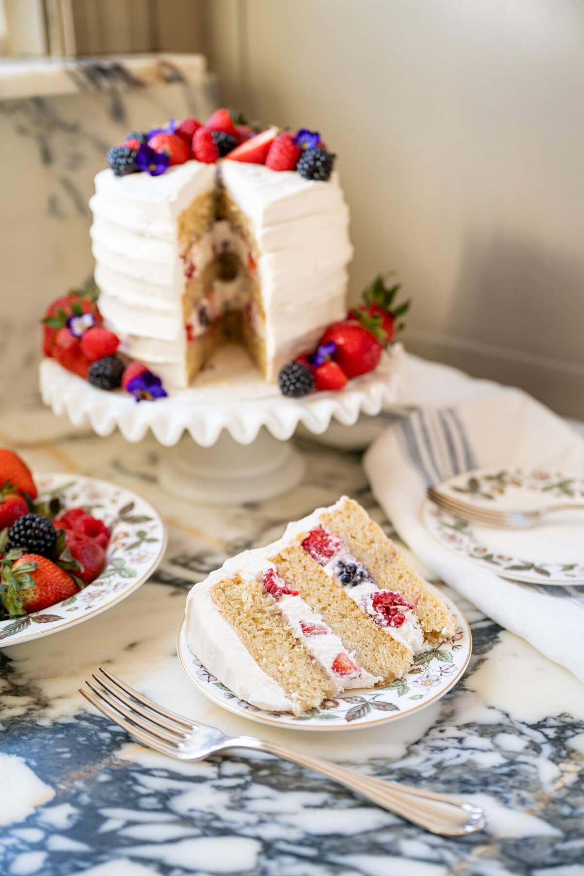 A layered vanilla cake with white frosting and fresh berries on top sits on a cake stand—this Whole Foods Berry Chantilly Cake copycat recipe features a slice filled with berries and cream, served on a floral plate, with more berries nearby on the marble counter.