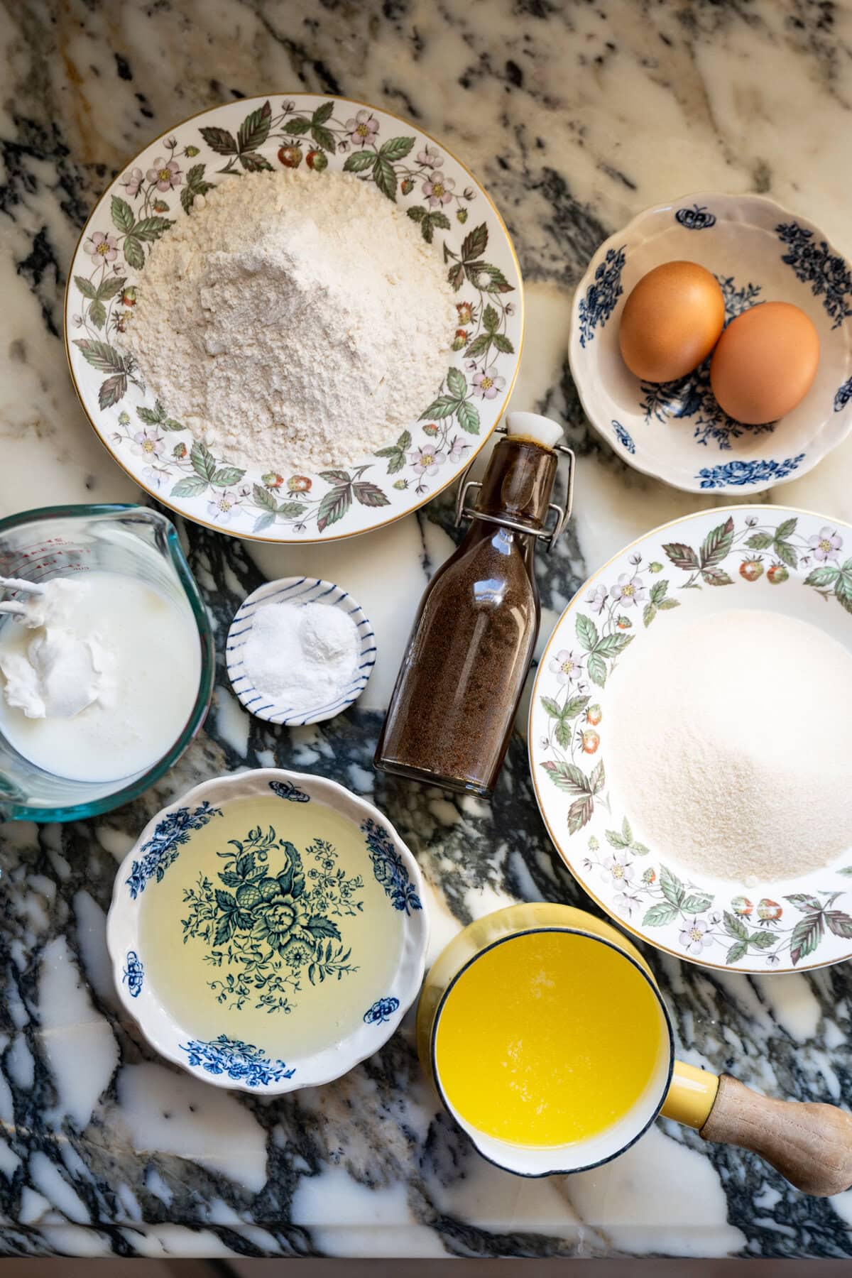 A marble countertop with bowls of flour, sugar, oil, melted butter, a small jug of buttermilk mixed with sour cream, two eggs, baking powder, and vanilla extract— berry chantilly cake ingredients arranged neatly.