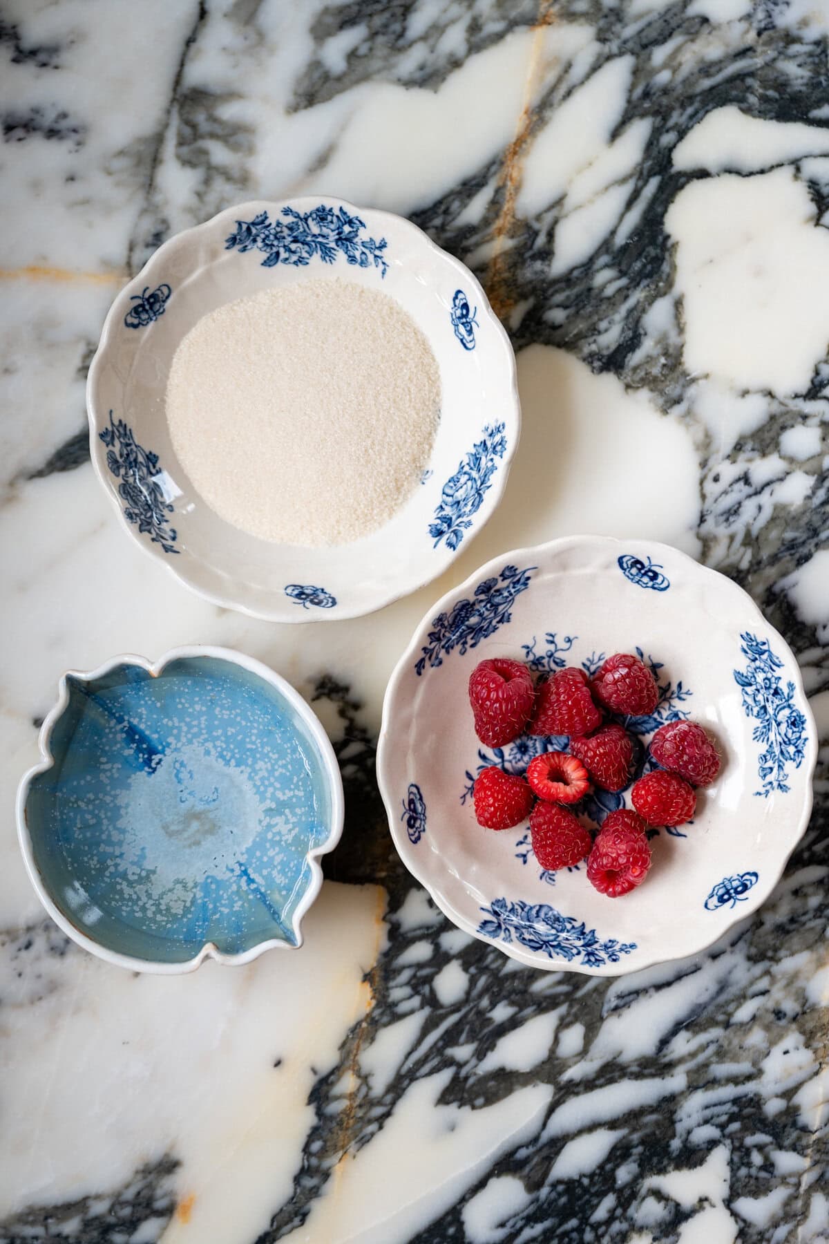 Three decorative bowls on a marble surface: one with granulated sugar, one with fresh raspberries, and one with water—stunning as raspberry simple syrup ingredients for a berry chantilly cake recipe.