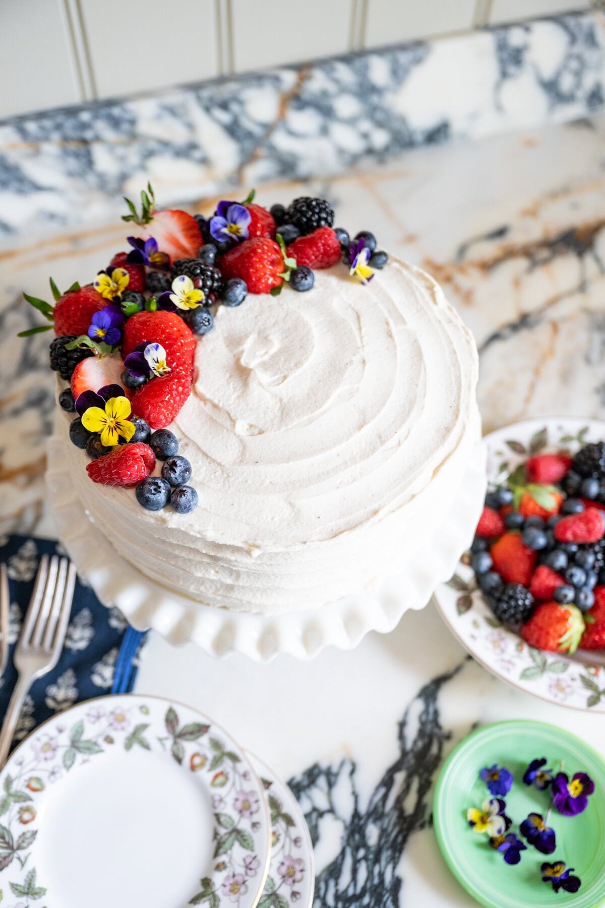 A white frosted cake, inspired by the Whole Foods Berry Chantilly Cake recipe, is topped with strawberries, blueberries, blackberries, and edible flowers. Plates, forks, and a small dish of extra berries rest nearby on a marble surface.