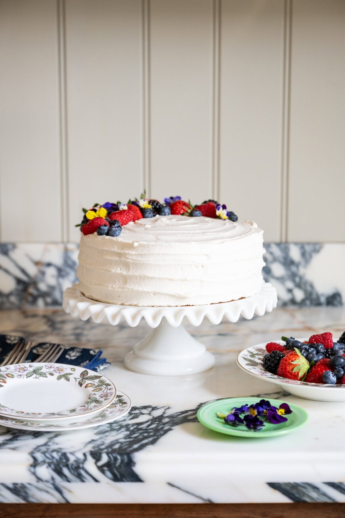 A frosted berry chantilly cake recipe is brought to life with assorted berries and edible flowers, elegantly arranged on a white cake stand atop a marble countertop, surrounded by plates and utensils.