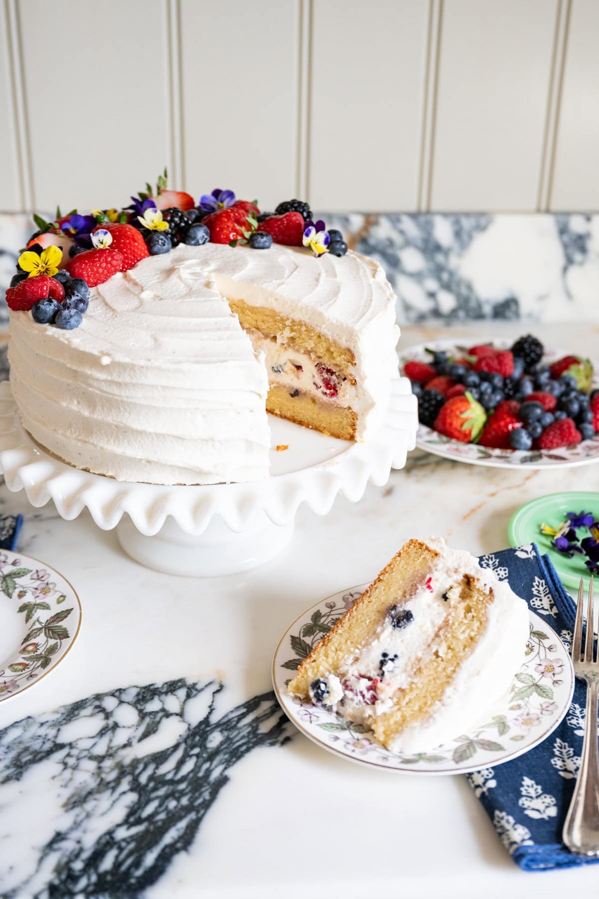 A frosted berry chantilly cake, decorated with berries and edible flowers, sits on a white ruffled cake stand. A slice is cut out to reveal the berry cream filling.