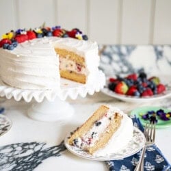 A layered vanilla cake with white frosting and berries on top sits on a cake stand. A slice with visible berries inside is on a plate nearby, next to a fork and blue napkin. More berries and flowers are in the background.