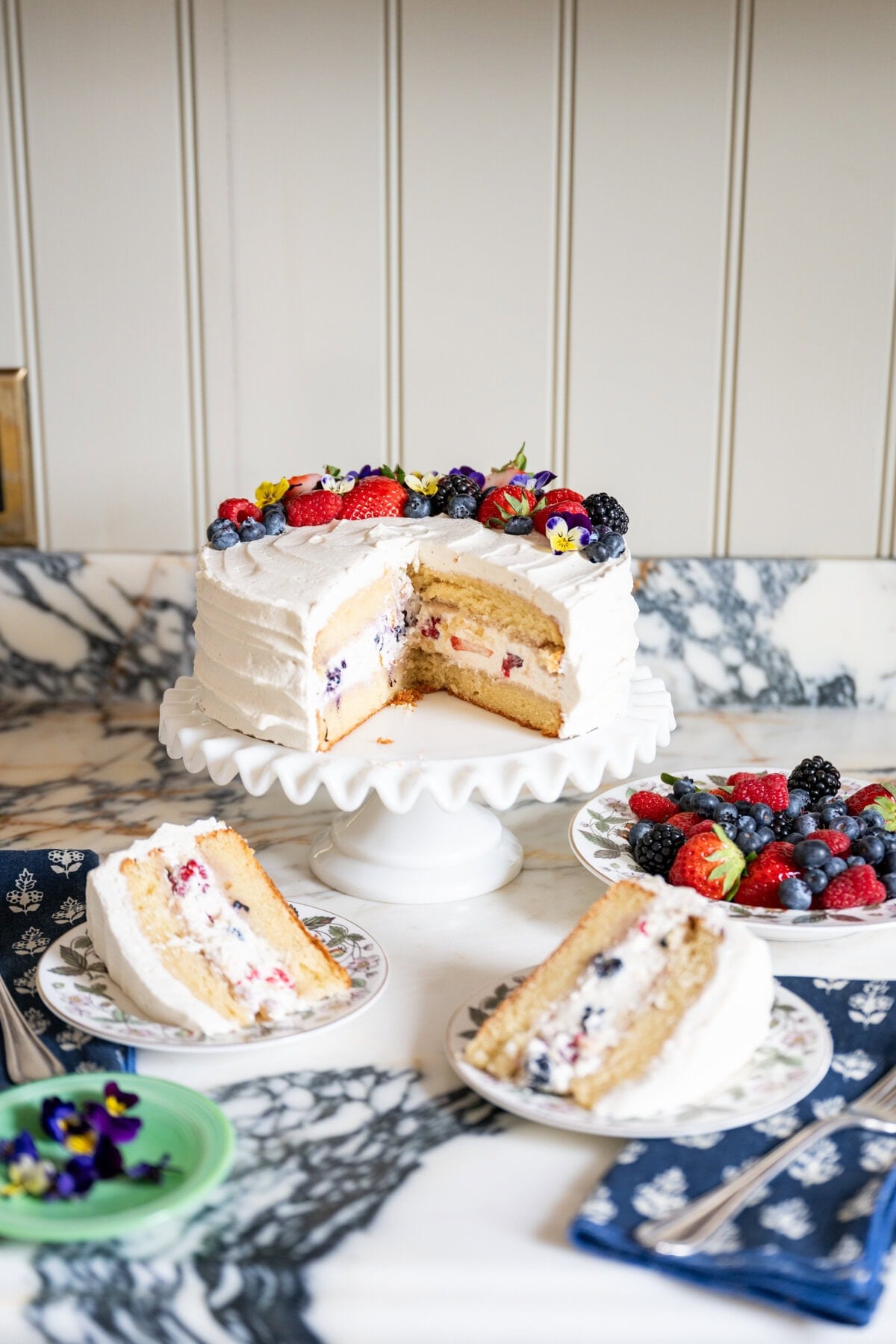 A white frosted berry chantilly cake with berries and edible flowers on top sits on a cake stand. Slices reveal a berry and cream filling, served with fresh berries on plates, all arranged on a marble countertop.