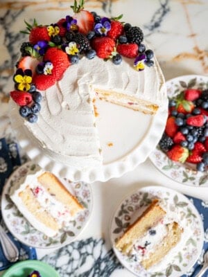 A white frosted berry chantilly cake sits on a cake stand, topped with fresh berries and edible flowers. Two slices rest on floral plates, alongside an extra plate of mixed berries on a marble surface.