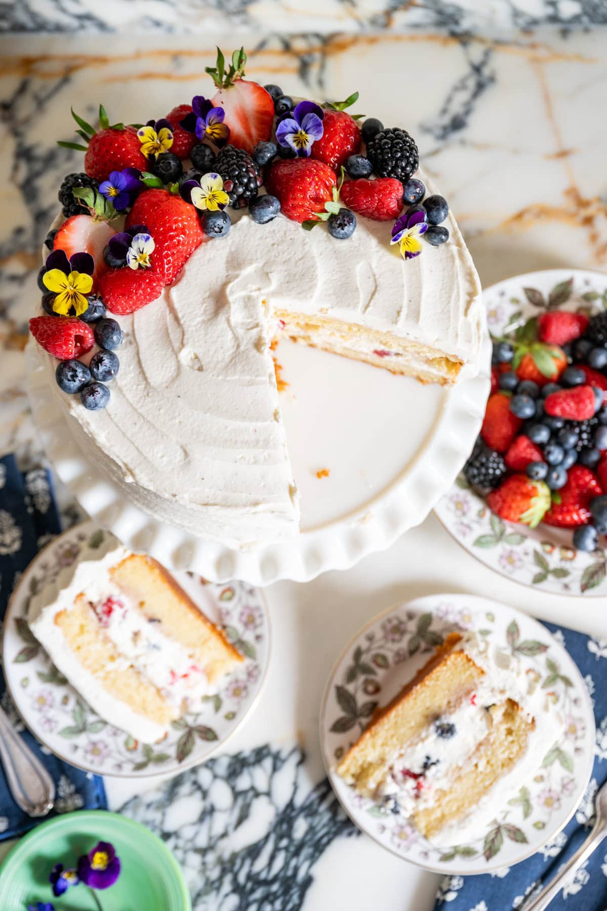 A white frosted berry chantilly cake sits on a cake stand, topped with fresh berries and edible flowers. Two slices rest on floral plates, alongside an extra plate of mixed berries on a marble surface.