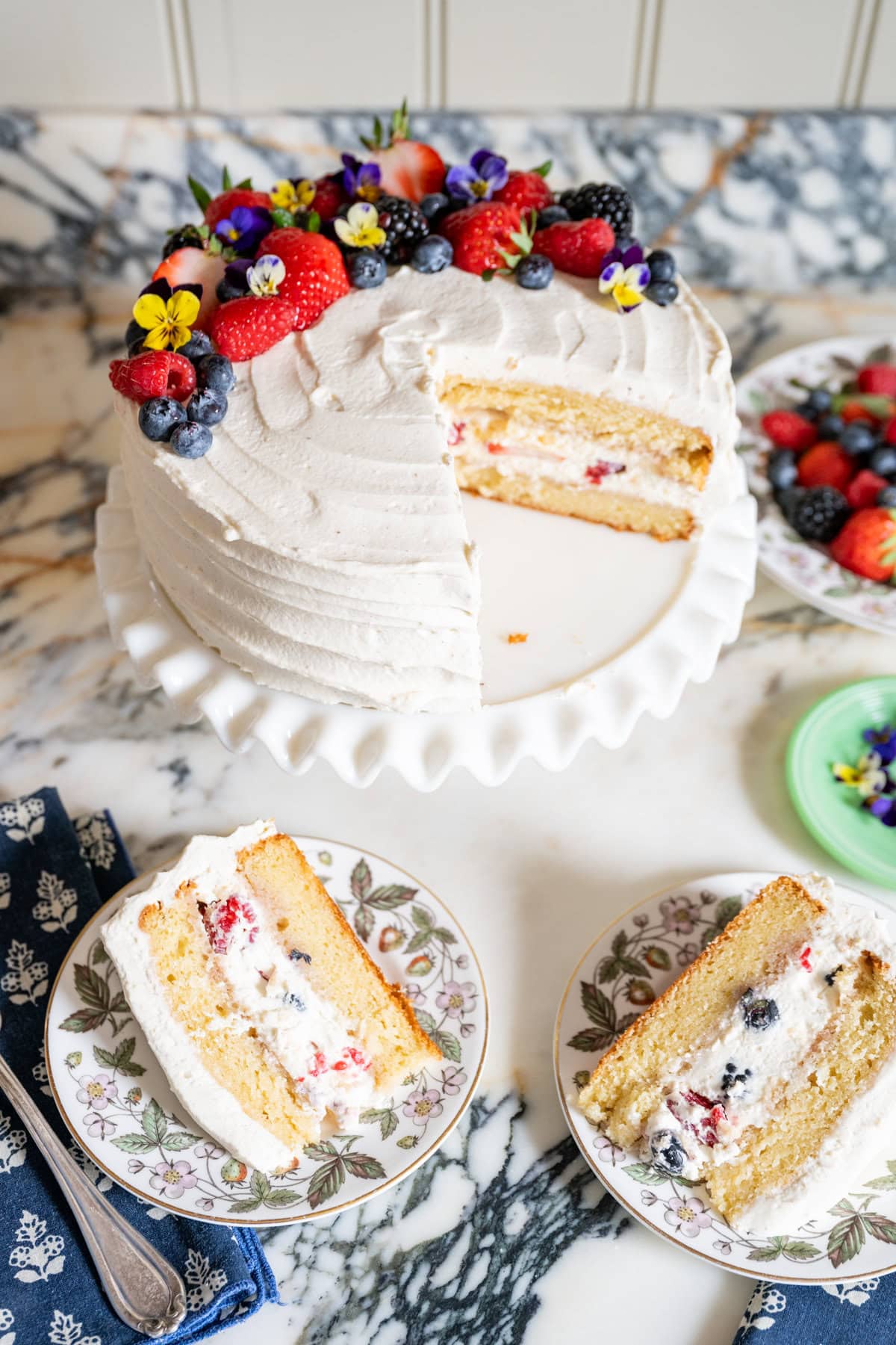 A berry chantilly cake topped with fresh berries and edible flowers sits on a marble surface. Two slices are served on floral plates, revealing the cake’s luscious, berry-filled interior cream layers.