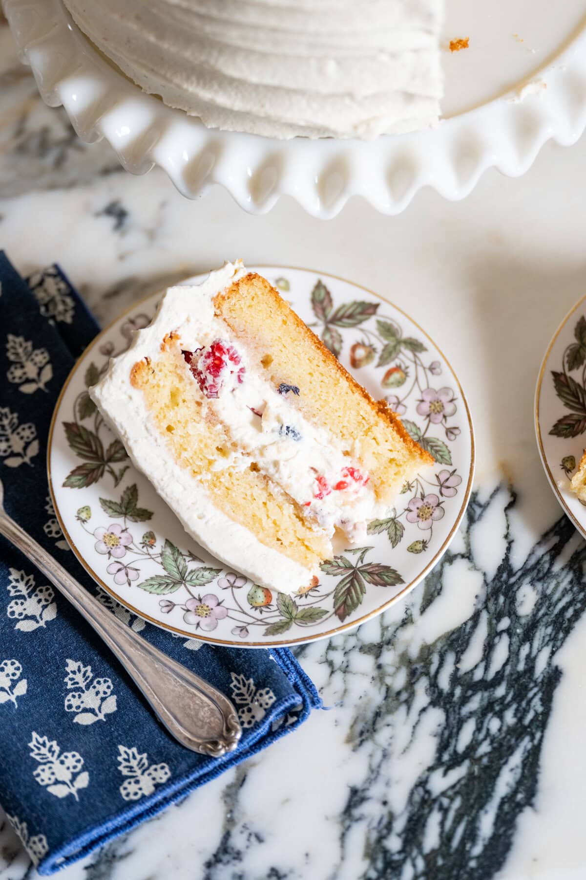 A slice of vanilla layer cake with whipped cream and berries filling sits on a floral-patterned plate beside a blue napkin and silver fork, with the rest of this berry chantilly cake recipe displayed on a white cake stand in the background.