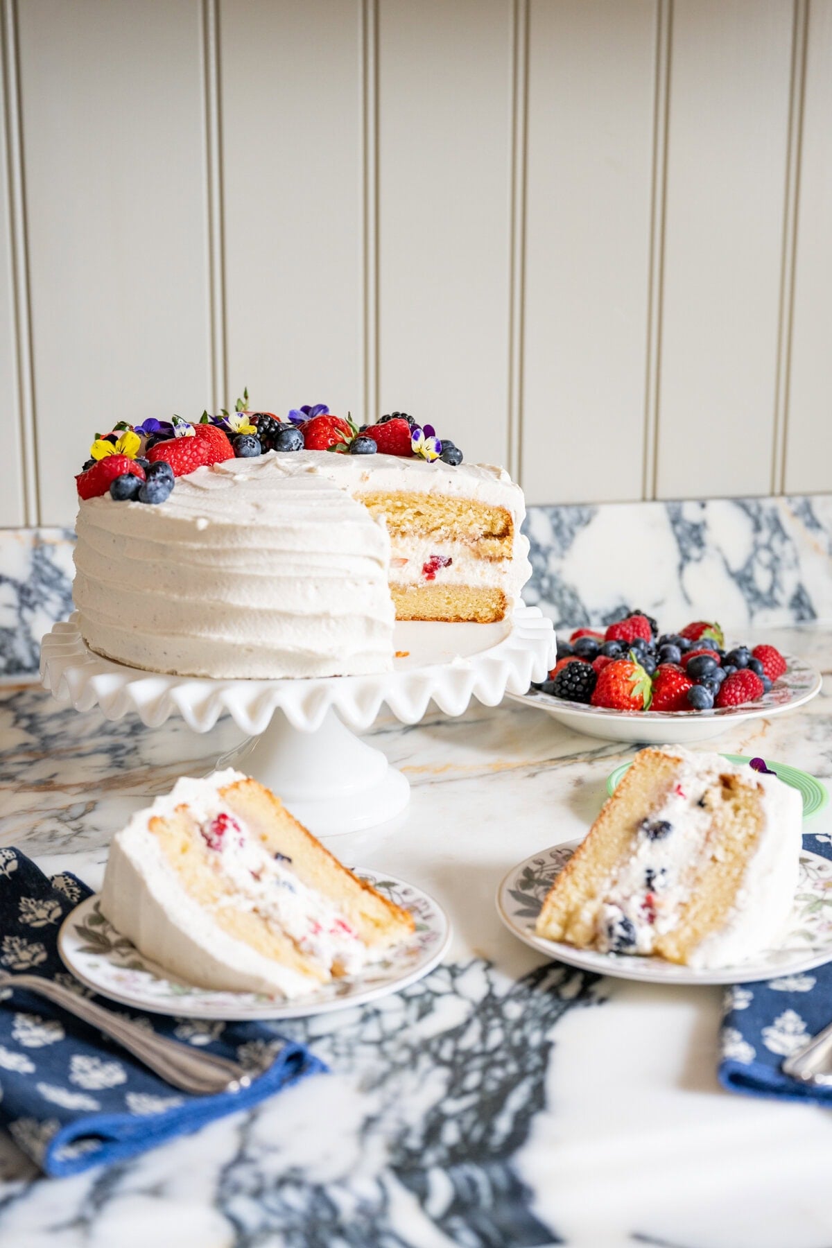 A frosted berry chantilly cake recipe masterpiece sits on a white cake stand, topped with fresh berries. Two slices on plates reveal colorful layers of cake and berries, while a bowl of mixed berries rests on the marble counter.
