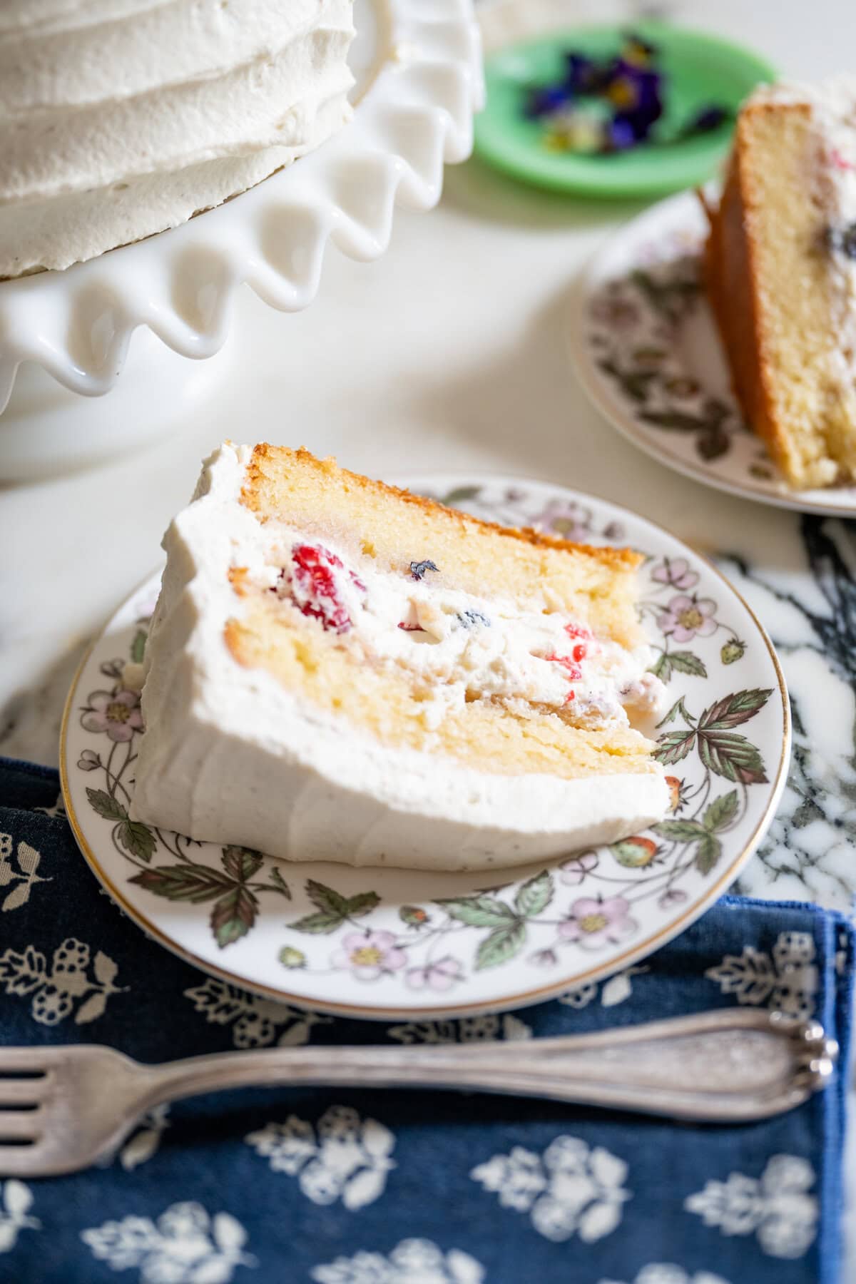 A slice of berry chantilly cake with whipped cream frosting and visible berries inside, served on a floral-patterned plate. The plate rests on a blue floral napkin with a fork beside it. The whole cake is in the background.