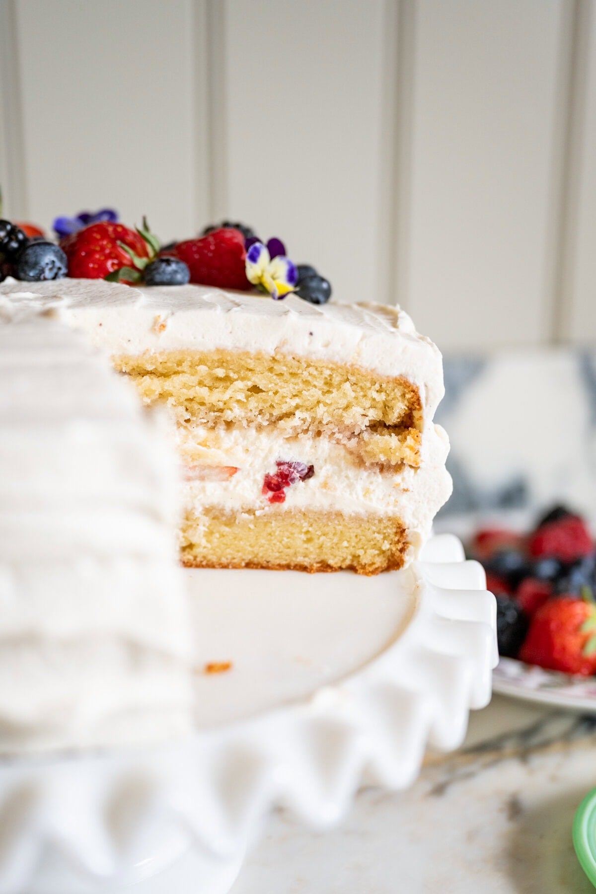 A layered vanilla berry chantilly cake recipe comes to life with white frosting, cream, and berries. Displayed on a white cake stand, it features assorted fresh berries and edible flowers atop, with a missing slice revealing its decadent inside.