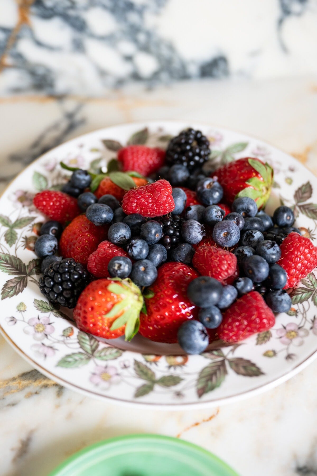 A decorative floral plate filled with fresh strawberries, raspberries, blackberries, and blueberries sits on a marble surface.