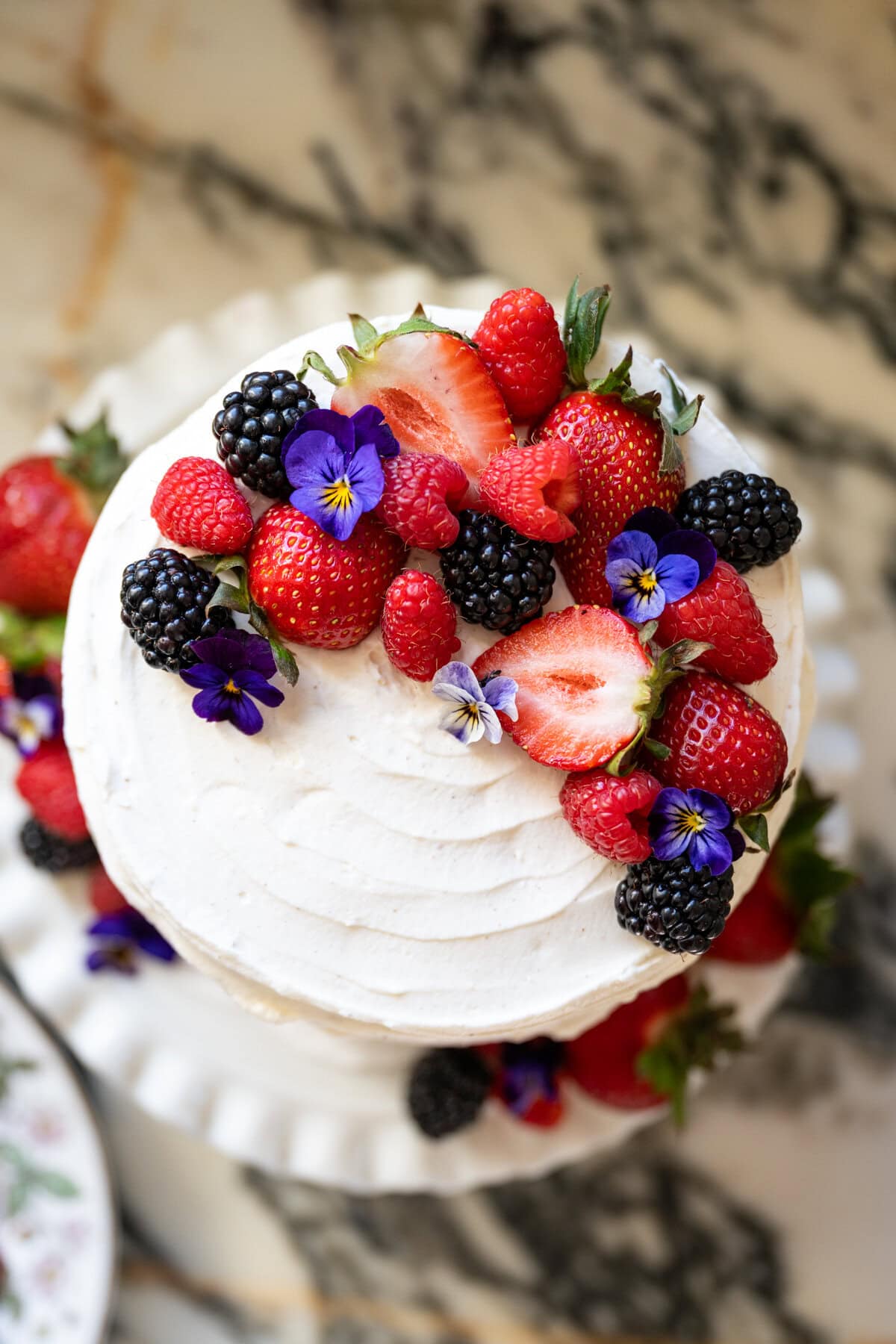 A white frosted cake inspired by the Whole Foods Berry Chantilly Cake is topped with fresh strawberries, raspberries, blackberries, and edible purple flowers, displayed on a white cake stand on a marble surface.