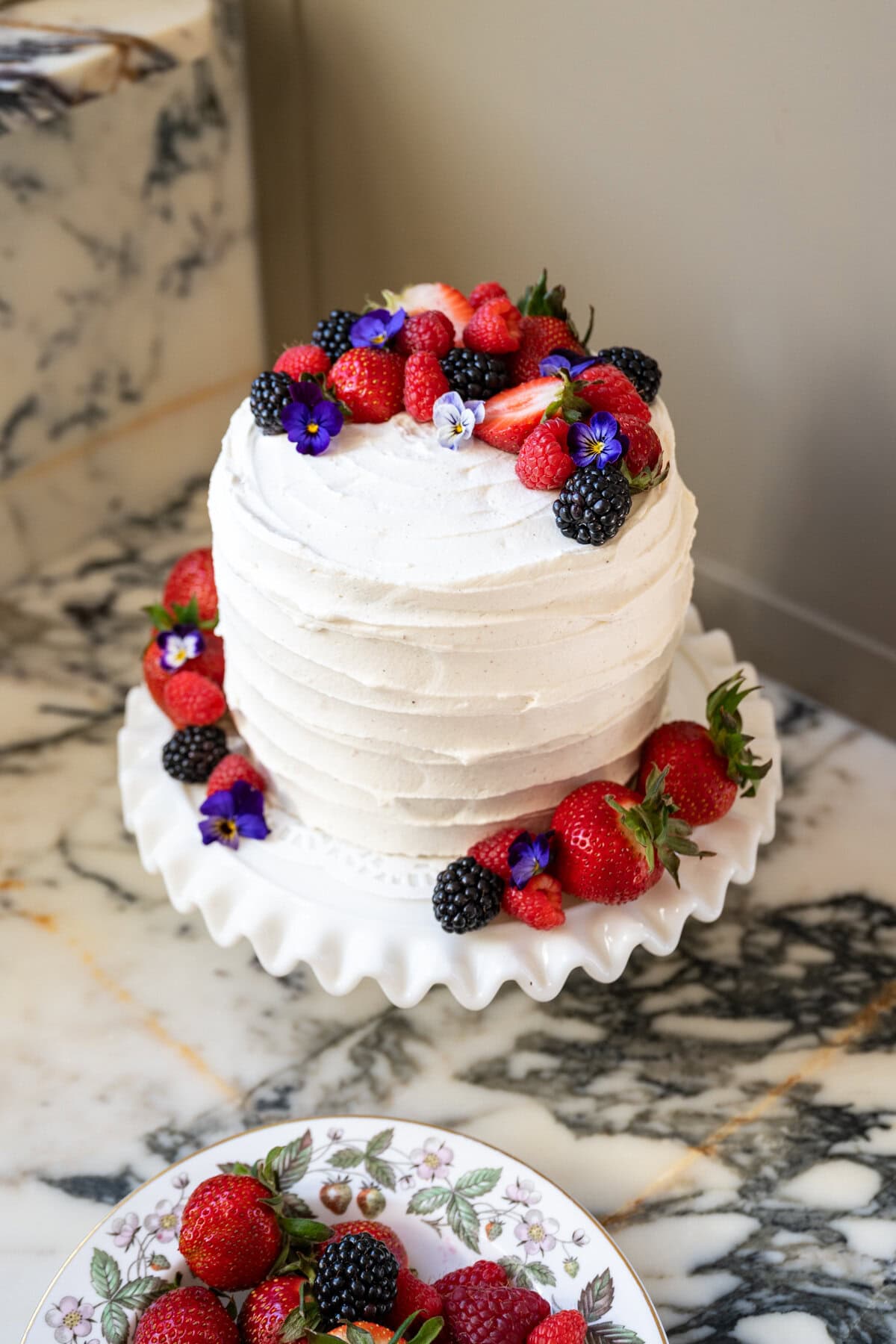 A white frosted layer cake on a ruffled cake stand, a Whole Food copycat berry chantilly cake recipe, is topped with strawberries, raspberries, blackberries, and edible purple flowers. Extra berries are scattered on a floral-patterned plate nearby.