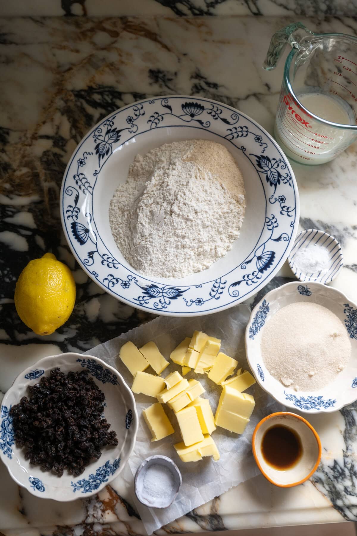 Irish soda bread scones recipe ingredients on a marble counter: a bowl of flour, a glass measuring cup with milk, a lemon, a bowl of currants, cubed butter, a small bowl of sugar, vanilla extract, and a pinch of salt.