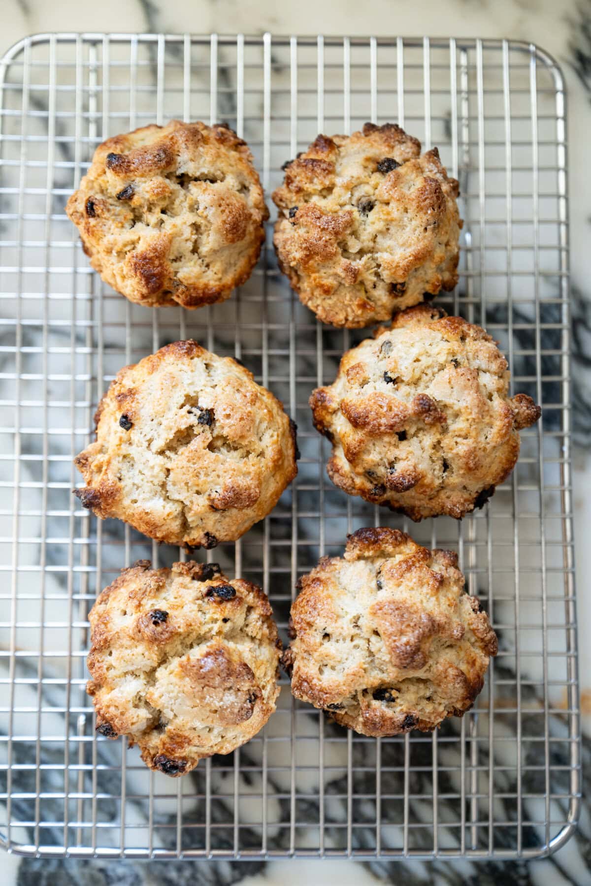 Six golden-brown Irish soda bread scones with a crisp, textured top are cooling on a wire rack placed over a marble surface. The scones have visible bits of currants, adding a classic touch to their delicious appearance.