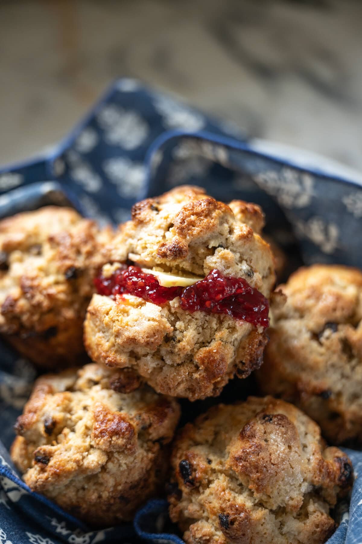A close-up of freshly baked Irish soda bread scones in a blue-patterned cloth. One scone, split open and filled with butter and jam, reveals its crumbly texture flecked with currants. The background is softly blurred.