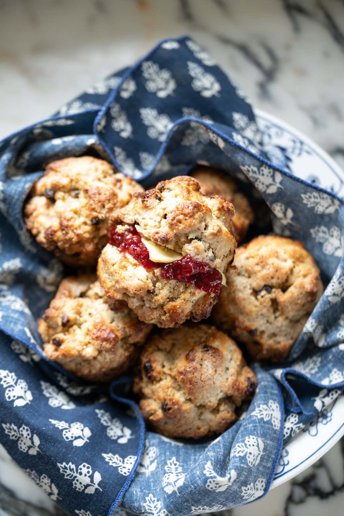 A basket lined with a blue and white floral cloth holds several golden brown scones made with Mary O's copycat recipe, one split open and filled with butter and jam. The basket is set on a marble surface.