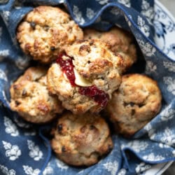 A bowl lined with blue and white patterned cloth holds several golden brown Irish soda bread scones, one split open and topped with butter and red jam, just like Mary O's copycat recipe—perfect with currants for a classic touch.